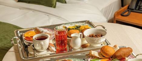 A neatly made bed with white and green pillows, a bedside table with a telephone, and a breakfast tray with various food items and drinks on a round table.