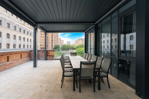 A covered outdoor dining area with a long table and chairs, large windows, and a view of surrounding buildings. - Hayasa Hotel Old Yerevan (Yerevan)