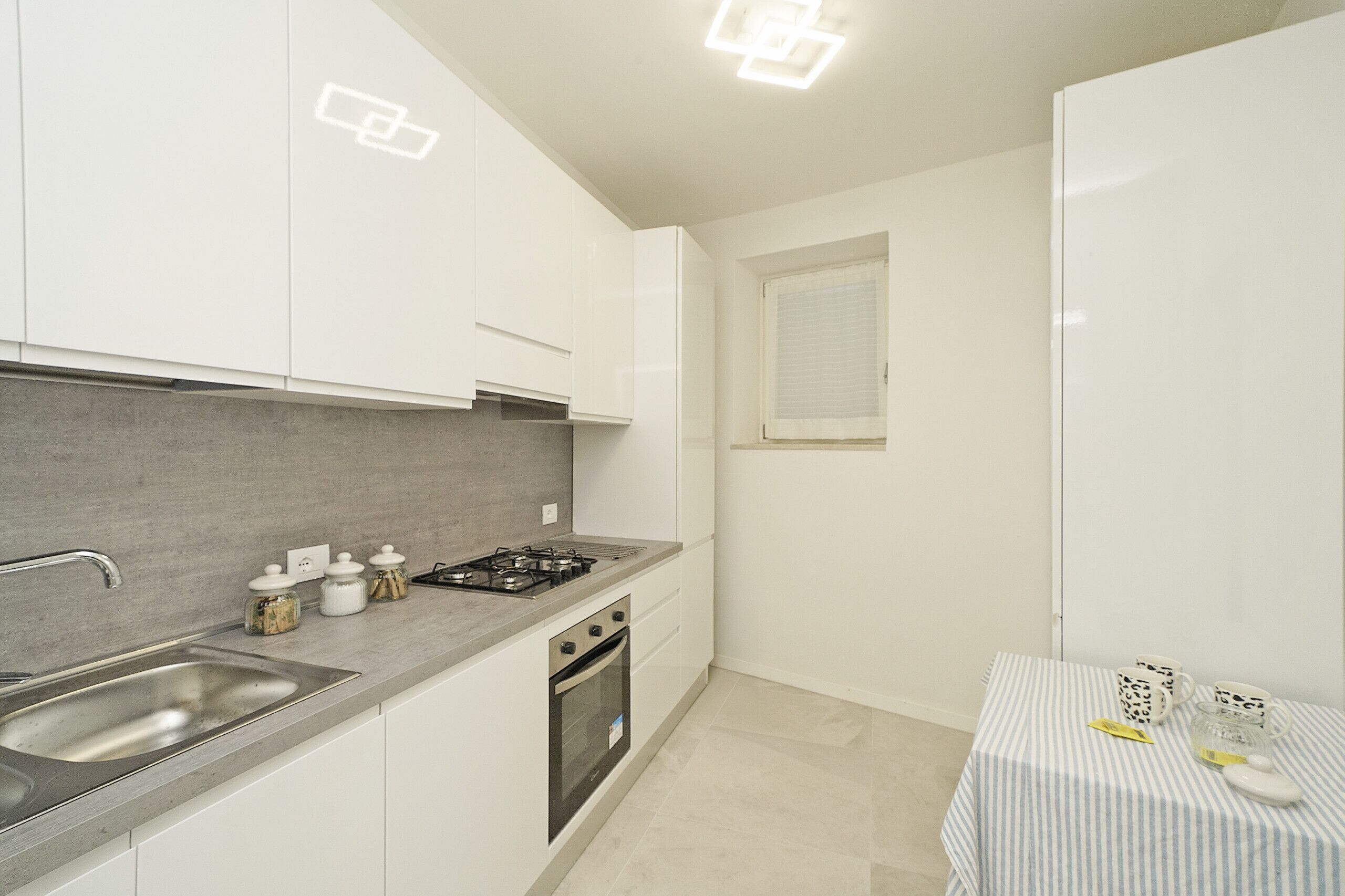 A compact kitchen with white cabinets, a grey countertop, a stainless steel sink, a gas stove, and a small window.