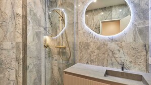 Un bagno moderno con parete in marmo, uno specchio rotondo, una doccia con accessori dorati e un mobile bagno in legno.