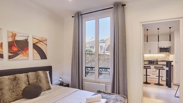 Une chambre d’hôtel moderne dotée d’un grand lit, offrant une vue sur la ville par une fenêtre, et comprenant une kitchenette dans la pièce adjacente.