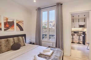 A modern hotel room with a large bed, a view of the city through a window, and a kitchenette in the adjacent room. - Charming apartment - Buttes Chaumont (Paris)
