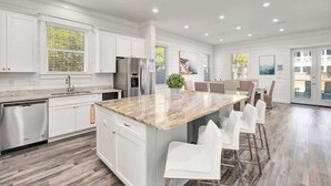 A modern kitchen with white cabinetry, a central island with bar stools, stainless steel appliances, and a large window with blinds. - Modern 3-story Beach House W Poll and Billiard (Miramar Beach)