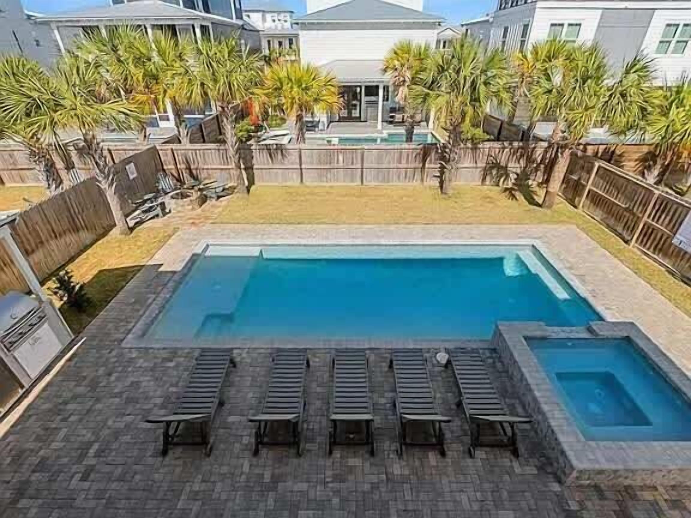 Piscina rectangular con solárium empedrado, rodeada de palmeras y una valla de madera.