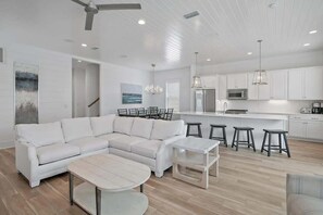 Un salon moderne avec un canapé d’angle blanc, une table basse ronde et un coin repas équipé de tabourets de bar.