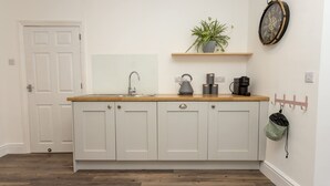 Una cocina con armarios blancos, encimera de madera, un lavabo, cafetera, hervidor, una planta en maceta y un reloj en la pared.