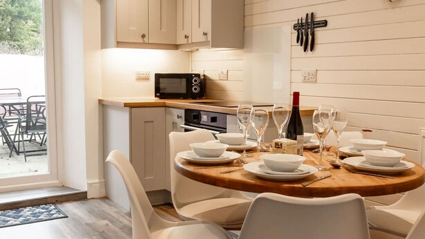 Una cocina moderna con armarios blancos, una mesa de madera para cuatro personas y vistas al exterior a través de una puerta de vidrio.