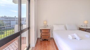 Habitación de hotel con balcón, cama con ropa blanca, mesita de noche de madera con lámpara y vistas a la ciudad.