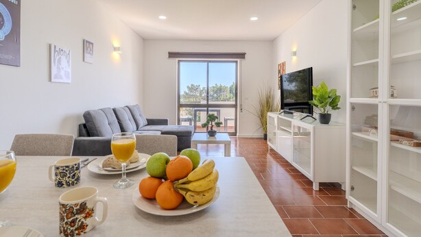 Una sala de estar moderna con un sofá, una mesa de comedor con frutas y bebidas, un televisor y un balcón con vistas.