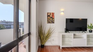 Una sala de estar moderna con un televisor de pantalla plana grande sobre un mueble blanco, una planta en maceta y un amplio ventanal con vistas a la ciudad.