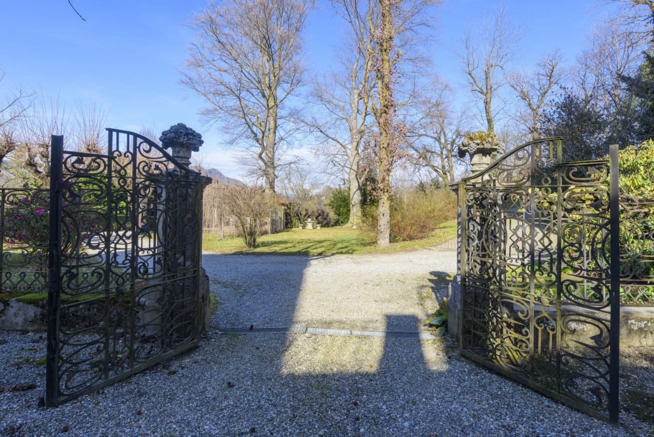 A gated entrance with ornate metalwork, a gravel path, and leafless trees in the background.