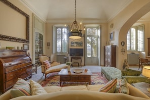 A spacious living room with a sofa, armchair, and a coffee table. There is a television, a fireplace, and a chandelier. - VARESE - Old-time villa next to golf course (Varese)