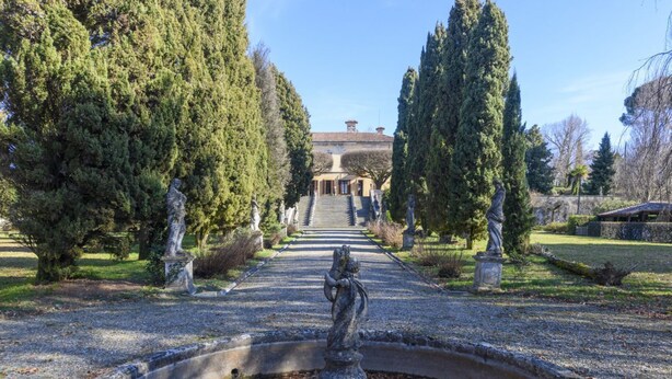 Ein Gartenweg führt zu einem Gebäude mit einer zentralen Statue und hohen Bäumen an beiden Seiten.