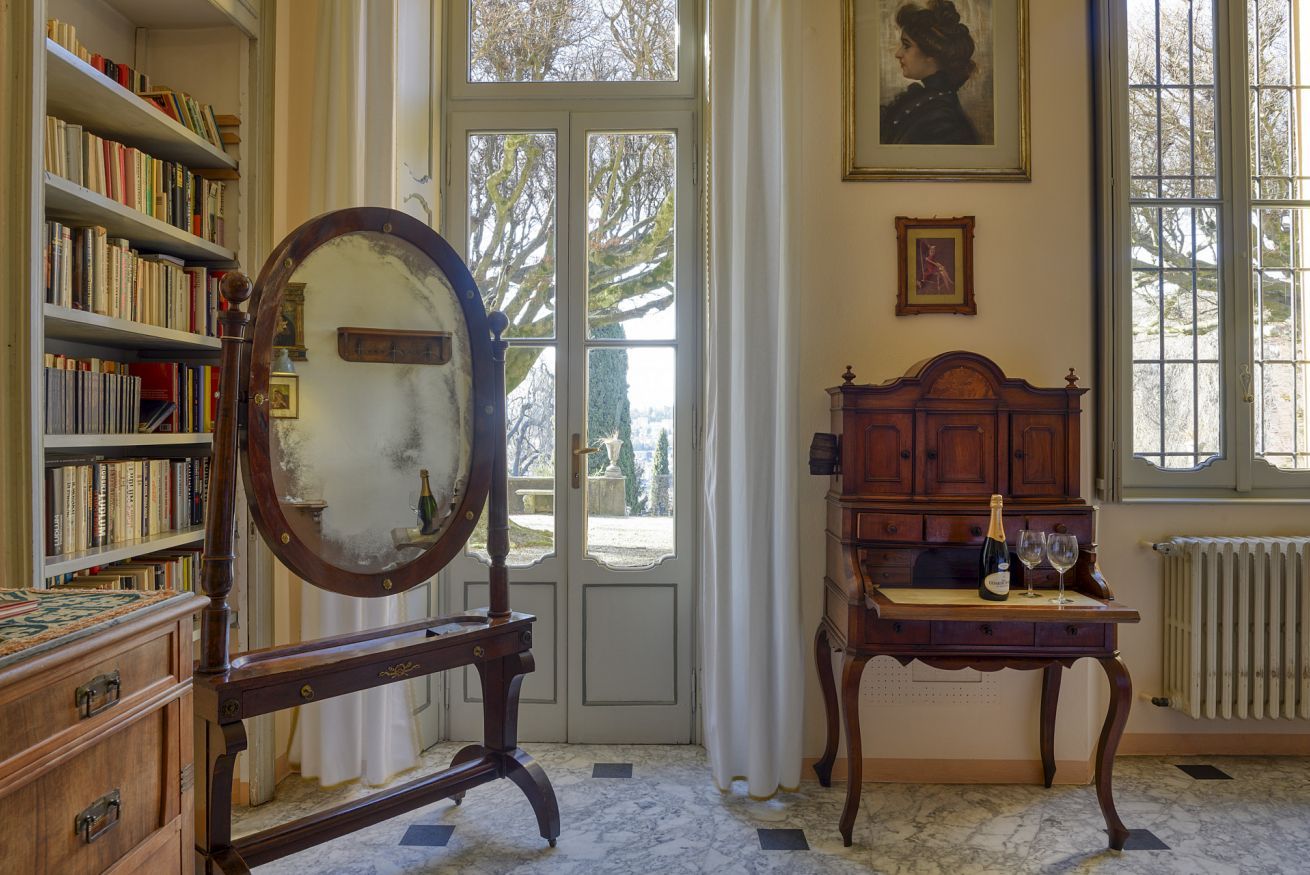 Een kamer met een grote spiegel, een houten kastje met een fles en glazen, een boekenkast, en een deur naar buiten.