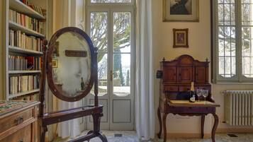 Een kamer met een grote spiegel, een houten kastje met een fles en glazen, een boekenkast, en een deur naar buiten.