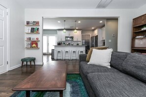 A modern living room with a grey sofa, a wooden coffee table, and a green ottoman. A kitchen with white cabinets and a bar area is visible in the background. - Stylish Cottage Near Stadium, BeltLine & Downtown (Atlanta)