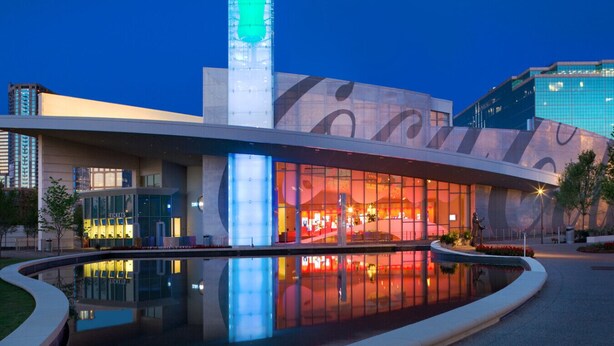 Ein modernes Gebäude mit einer großen Glasfassade, einer zentralen vertikalen Struktur und einem spiegelnden Wasserspiel davor.