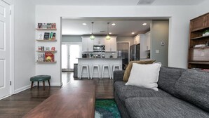 Ein modernes Wohnzimmer mit einem grauen Sofa, einem hölzernen Couchtisch und einem grünen Hocker. Im Hintergrund ist eine Küche mit weißen Schränken und einem Barhocker zu sehen.