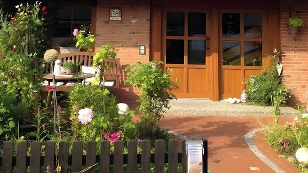 Une maison avec un toit en tuiles, une porte d’entrée en bois et un petit jardin agrémenté d’une clôture en bois et de plantes en pot.