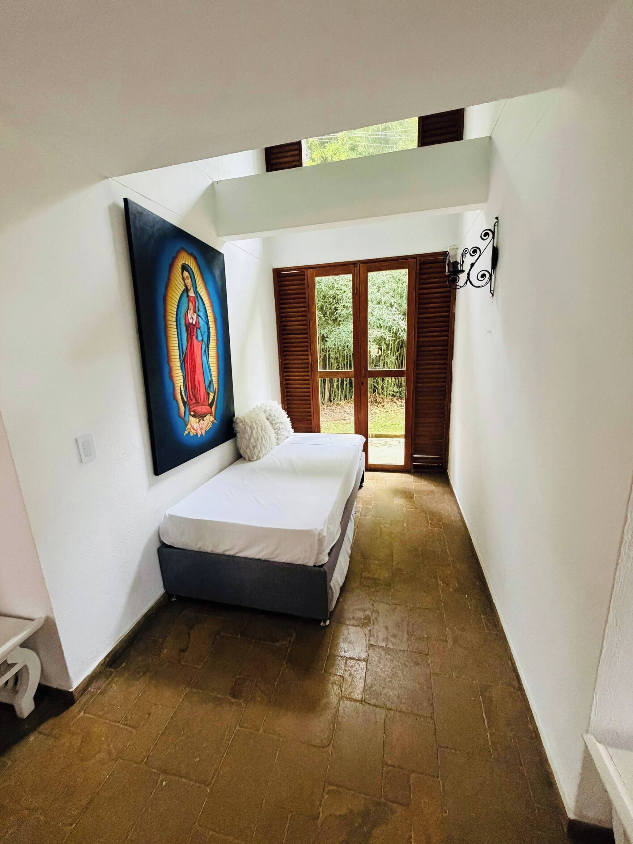 A bedroom with a bed, a painting, a window with shutters, and a door leading to an outdoor area.