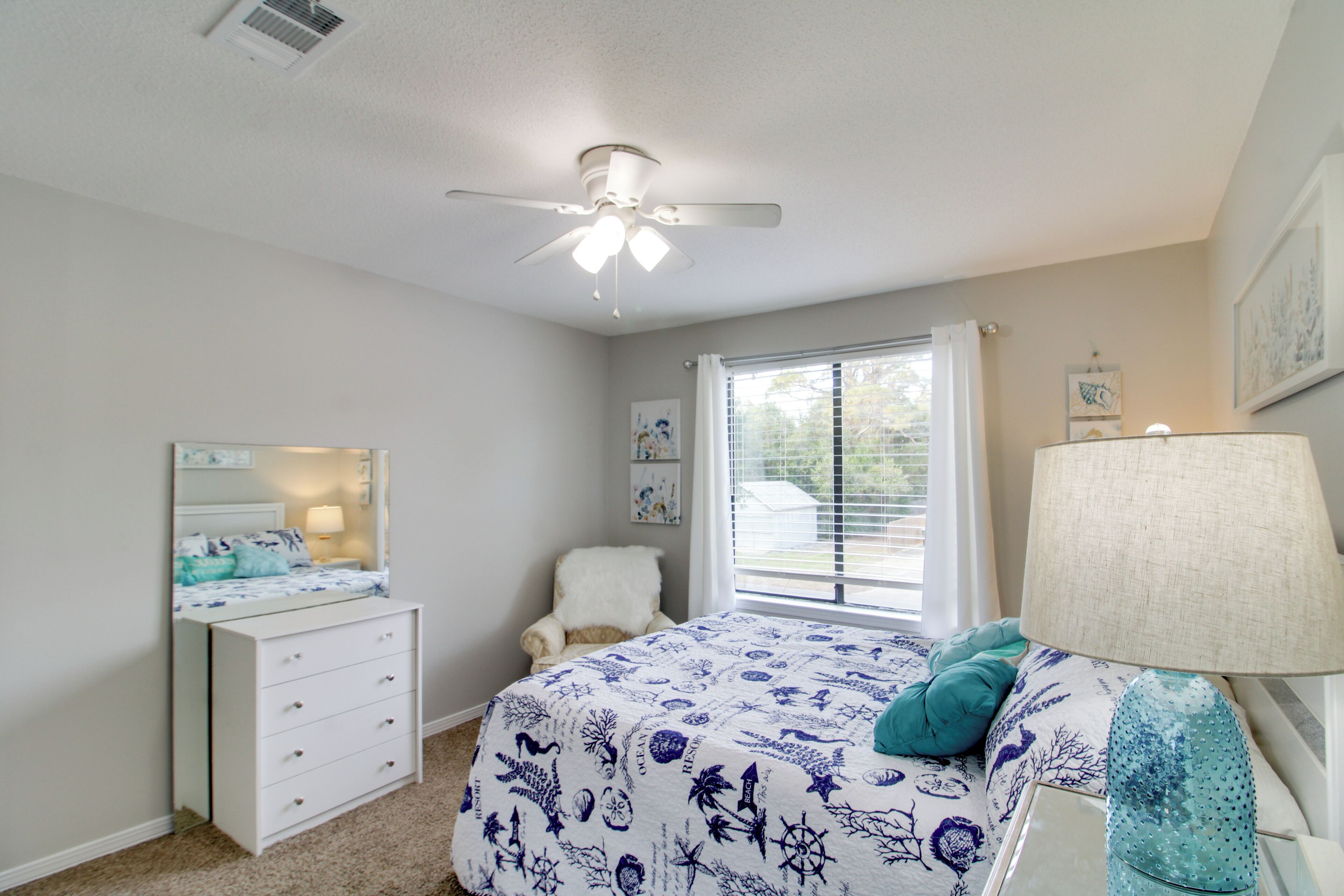 A bedroom with a bed, dresser, chair, ceiling fan, window, and lamp.