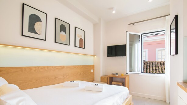 Habitación de hotel con cabecera de madera, cama con ropa blanca, televisor de pantalla plana en la pared y ventana con vista a un edificio.