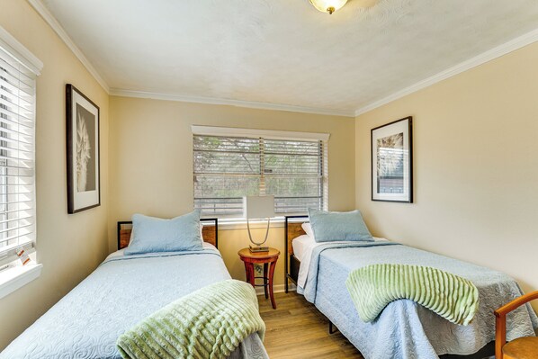A bedroom with two beds, a window with blinds, a bedside table, and a lamp.