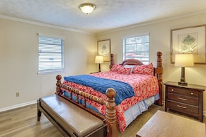 Un dormitorio con una cama de madera, un banco, dos mesitas de noche con lámparas y dos ventanas con persianas.