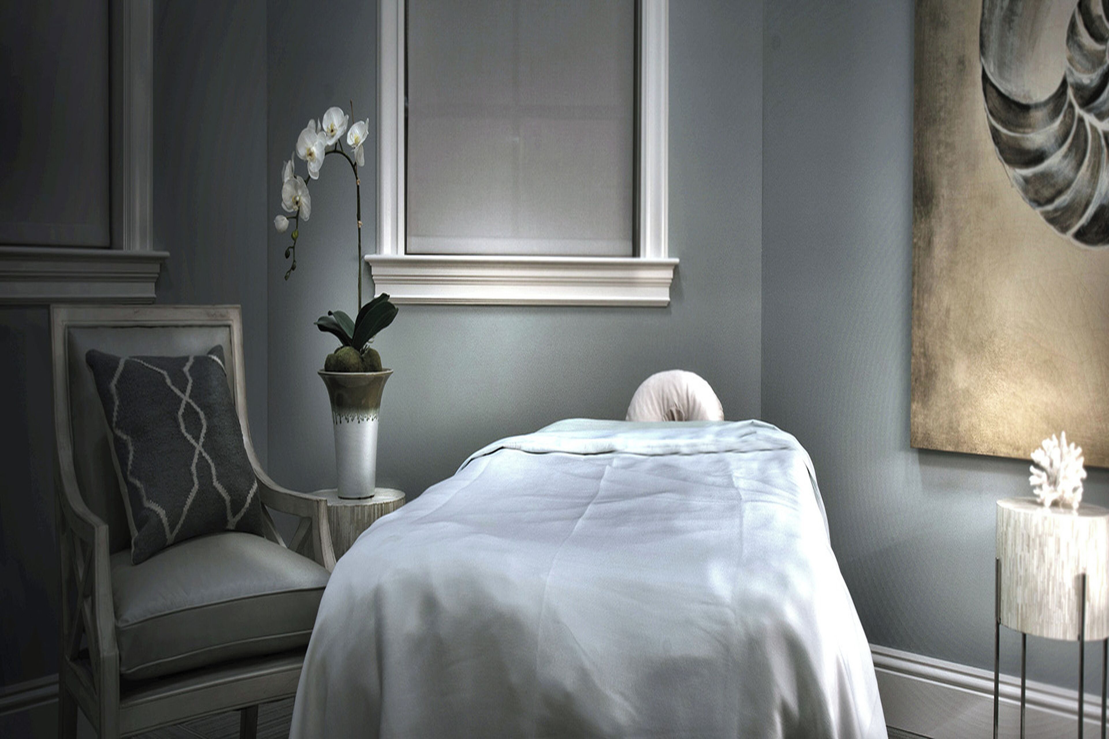 Une chambre à coucher avec un lit, une chaise, un vase de fleurs et un tableau accroché au mur.