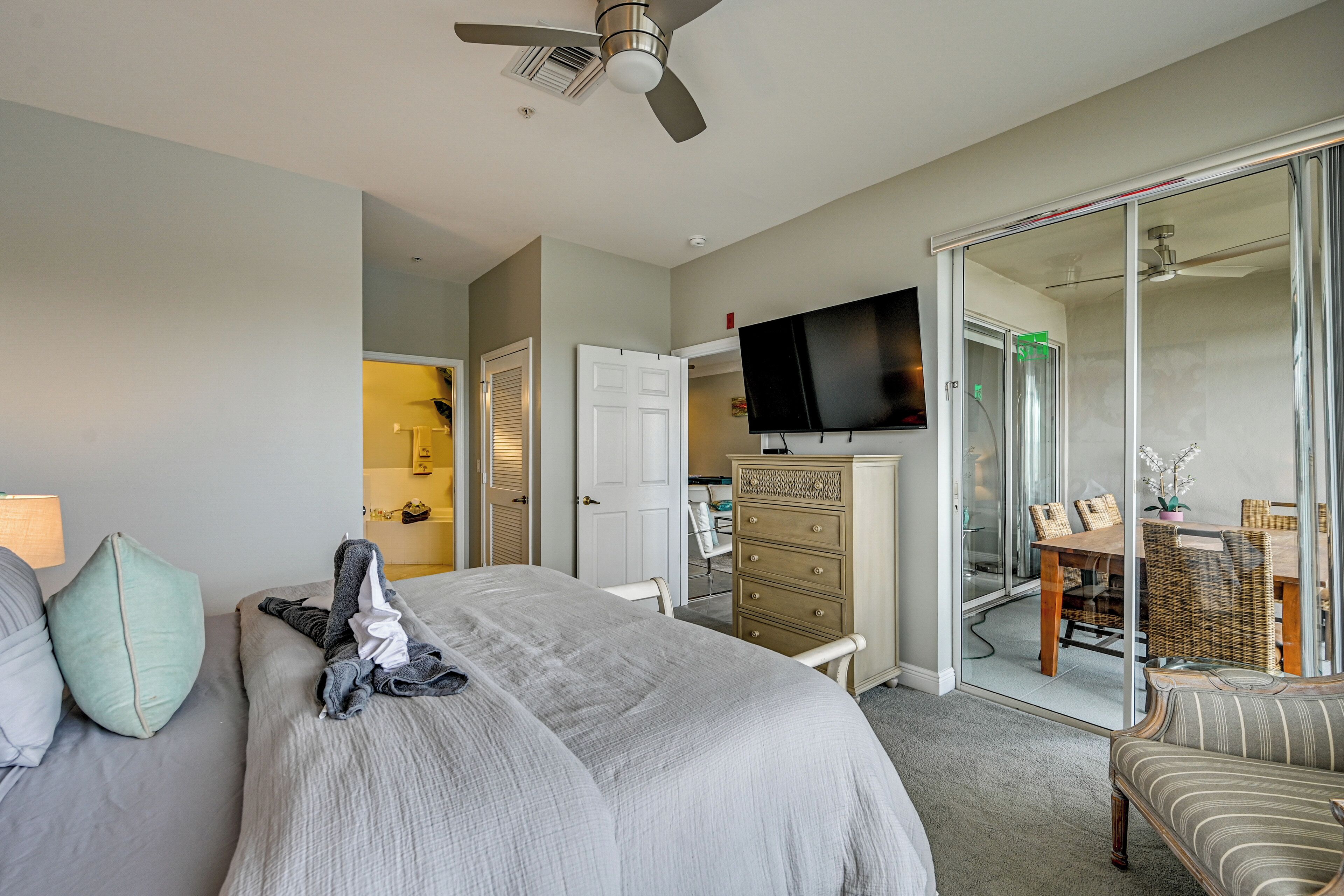 A hotel room with a bed, a dresser, a television, a dining area with a table and chairs, and a sliding glass door leading to a balcony.