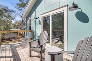 Een terras met Adirondack-stoelen en een tafel, met uitzicht op een huis met grote ramen en een lichtblauwe gevel.
