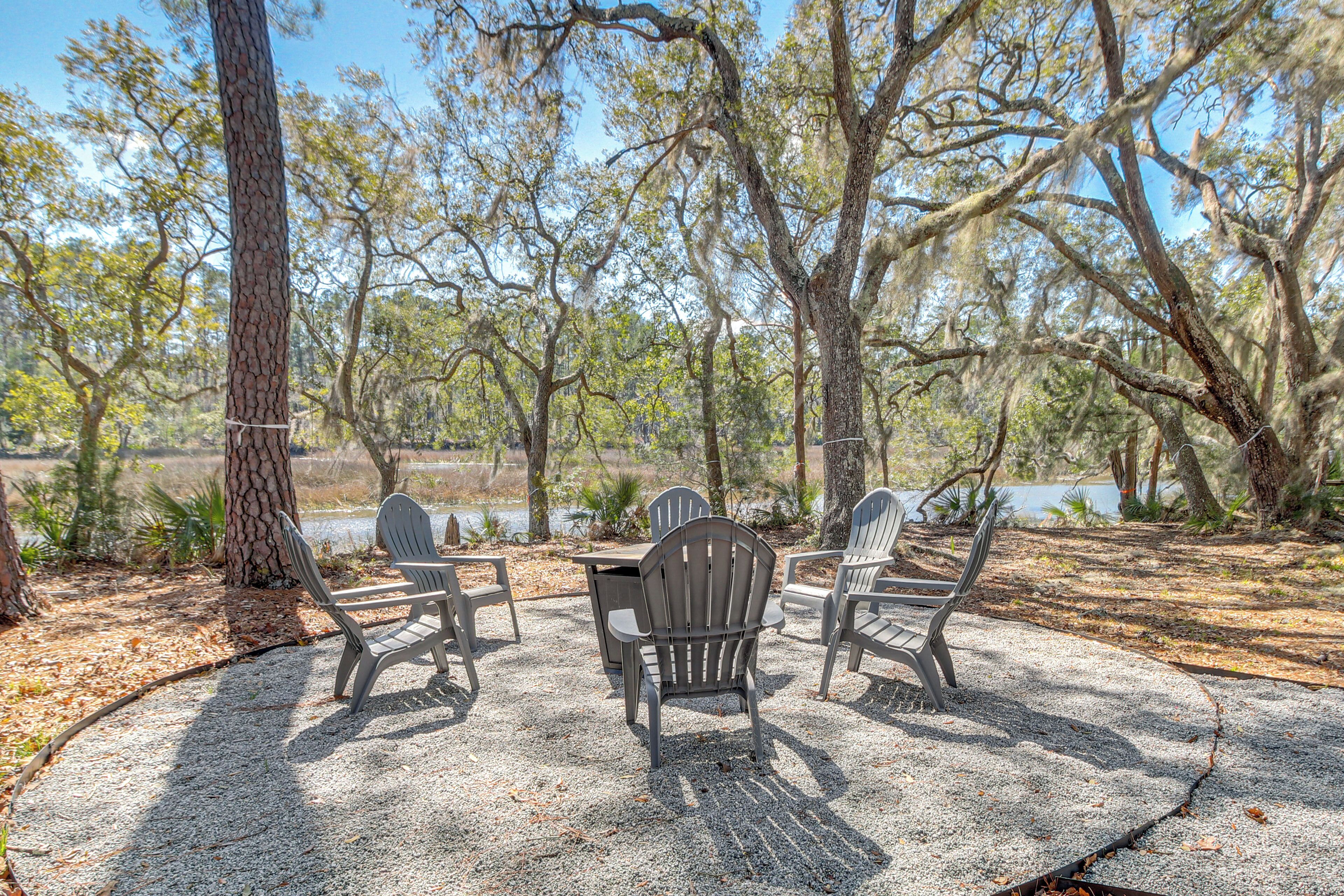 An outdoor seating area with Adirondack chairs and a table, surrounded by trees and a body of water.