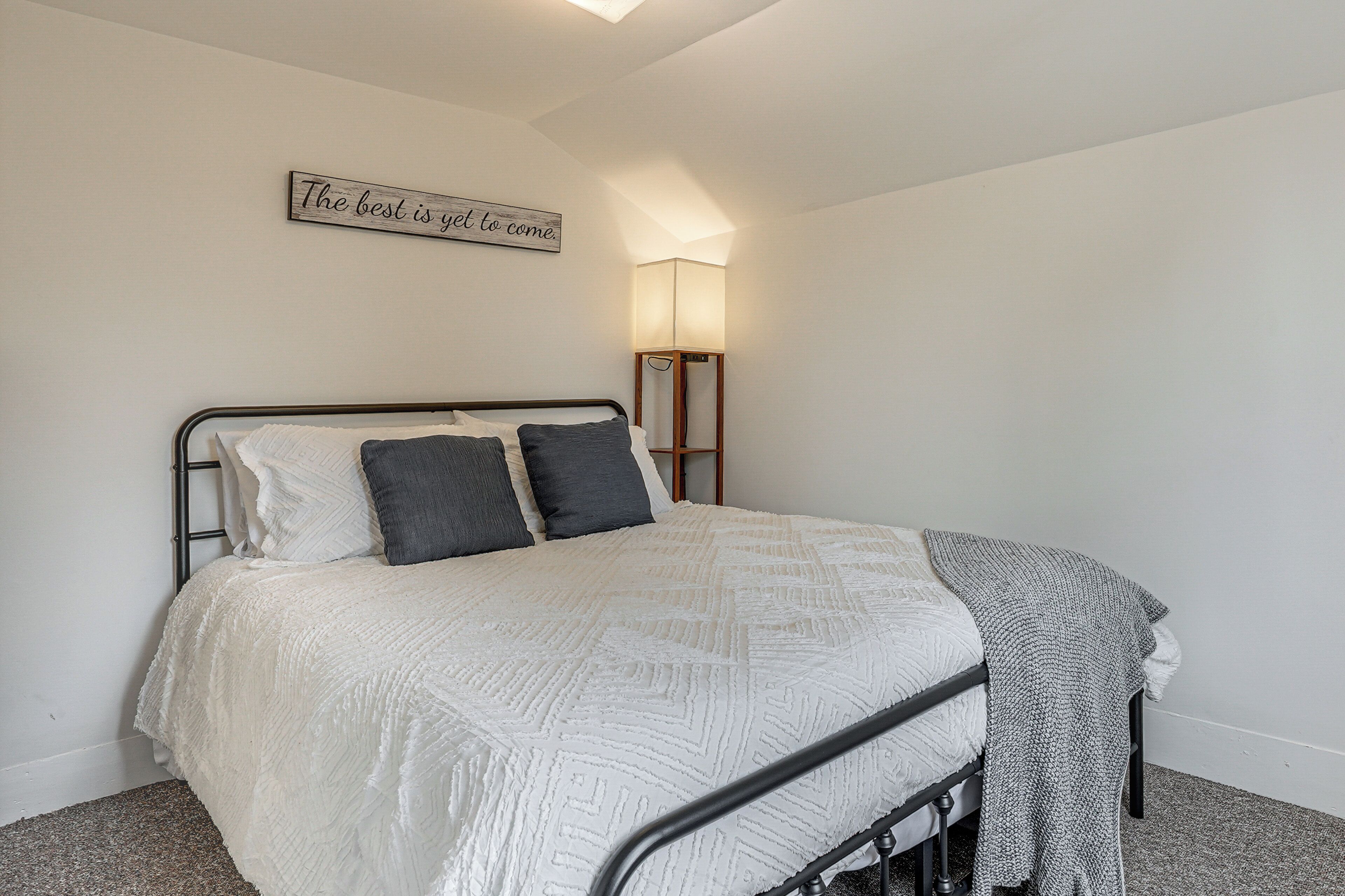 A neatly made bed with a white quilt and two pillows, a bedside lamp, and a wall sign.