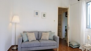 Een woonkamer met een grijze bank, een vloerlamp en een klein tafeltje met een kaars. Twee ingekaderde foto's hangen aan de muur en een deur leidt naar een andere kamer.