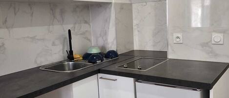 Una cocina compacta con gabinetes blancos, encimera negra, un fregadero, estufa y lavavajillas. Hay dos cubos de basura y un suelo de baldosas con diseño.