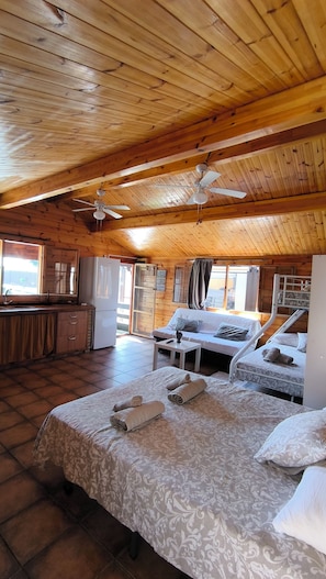 A cozy cabin-style room with a wooden ceiling, a bed with a patterned bedspread, a sofa, a small table, and a kitchenette area. - Villa Golondrinas Chipiona (AN)