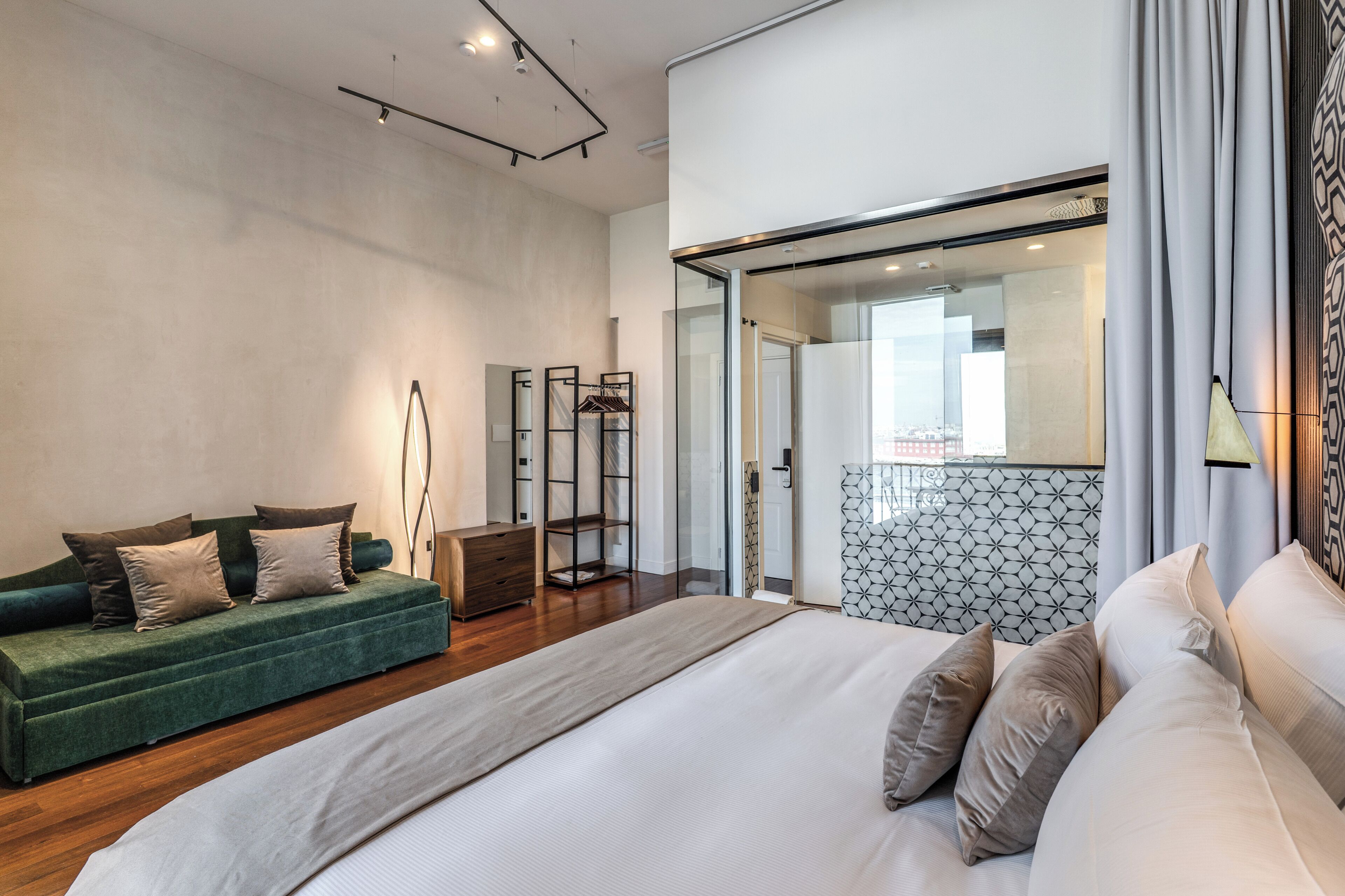 A modern hotel room with a large bed, a green sofa, a wooden dresser, and a glass door leading to a balcony.