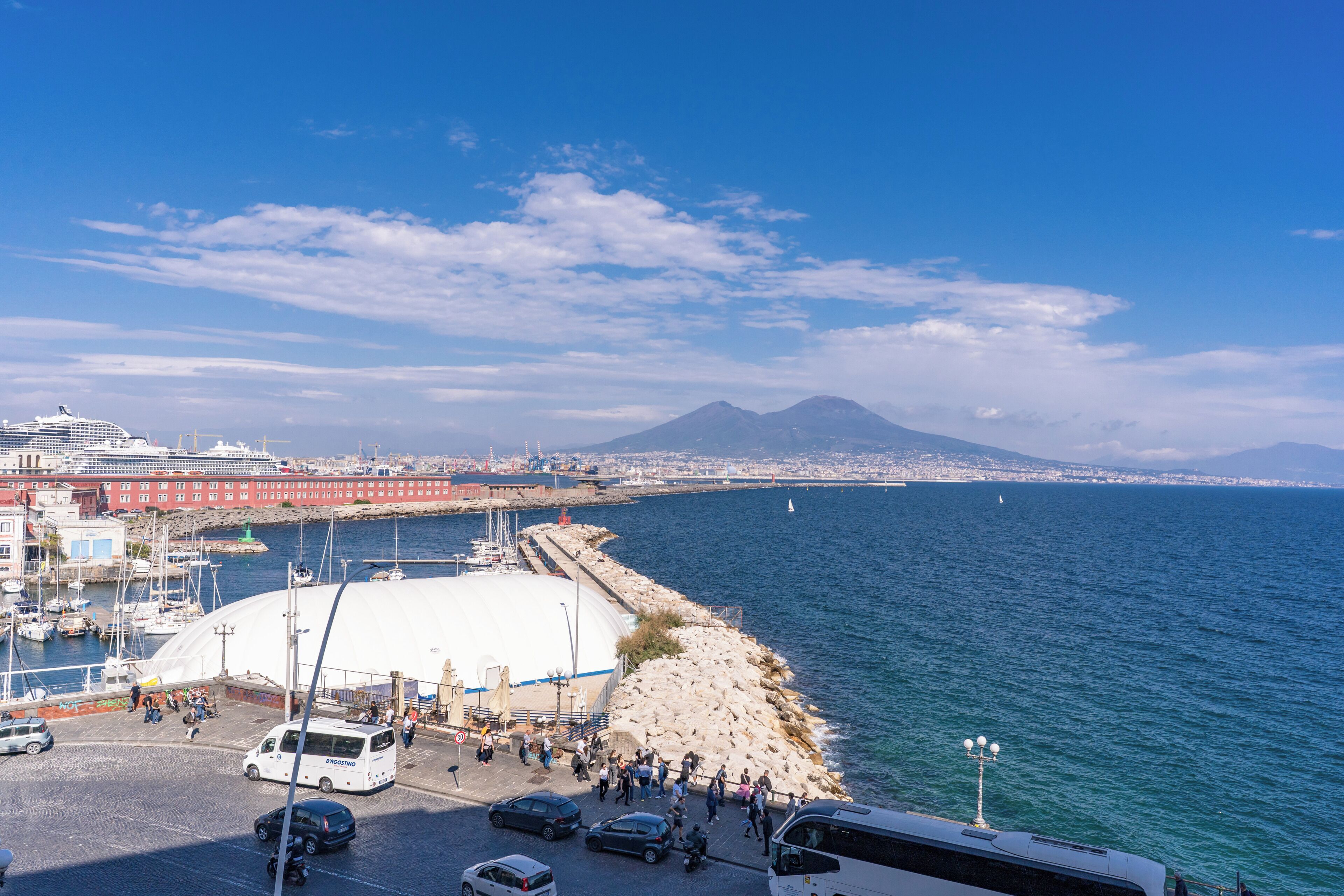 A coastal cityscape with a marina, a large white dome structure, and a mountain in the background.