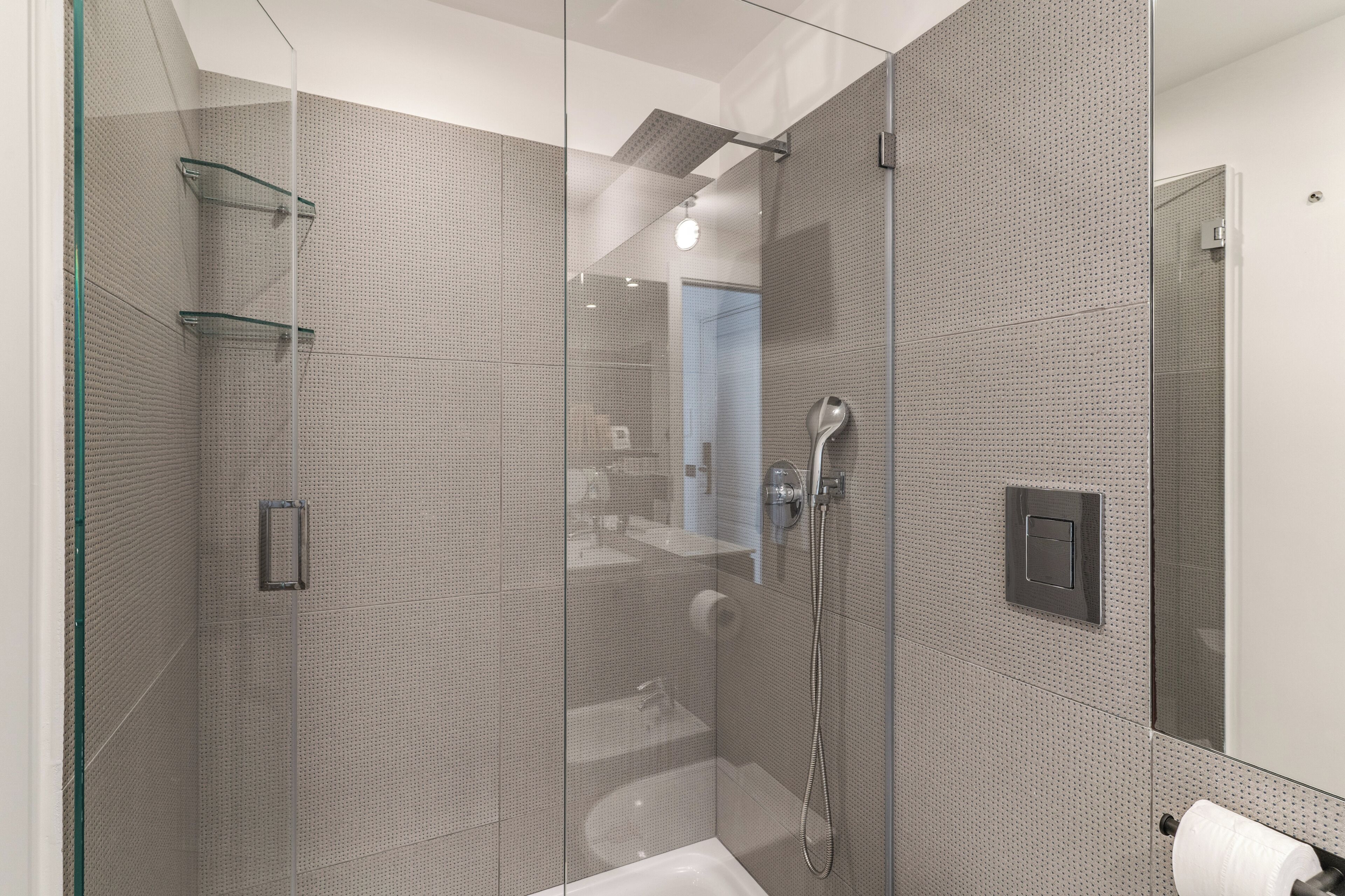 Une douche moderne dotée d’un pare-douche en verre, d’un pommeau de douche à main et d’un panneau de commande fixé au mur.