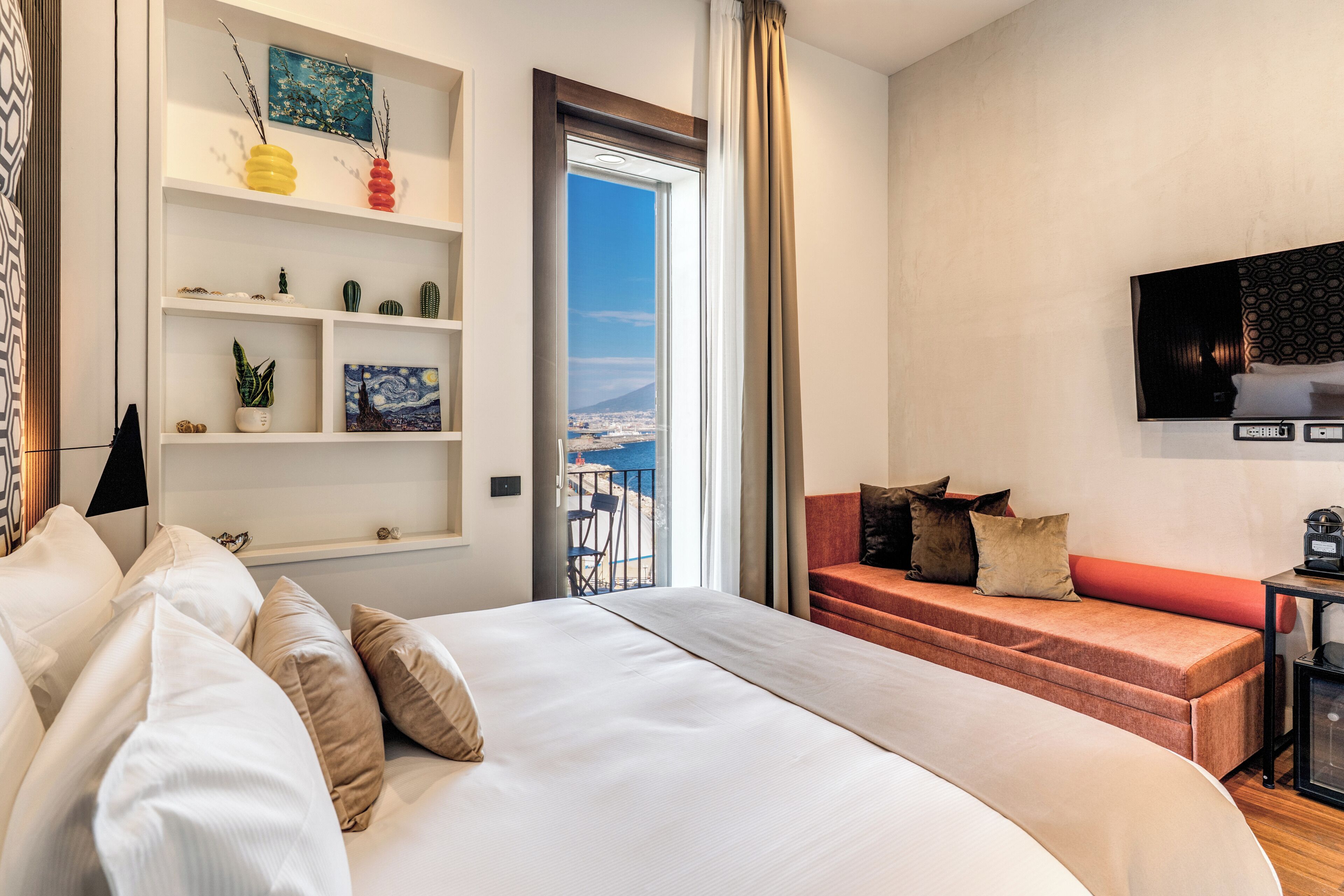 A hotel room with a bed, a sofa, a TV, a shelf with decorative items, and a view of the outside.