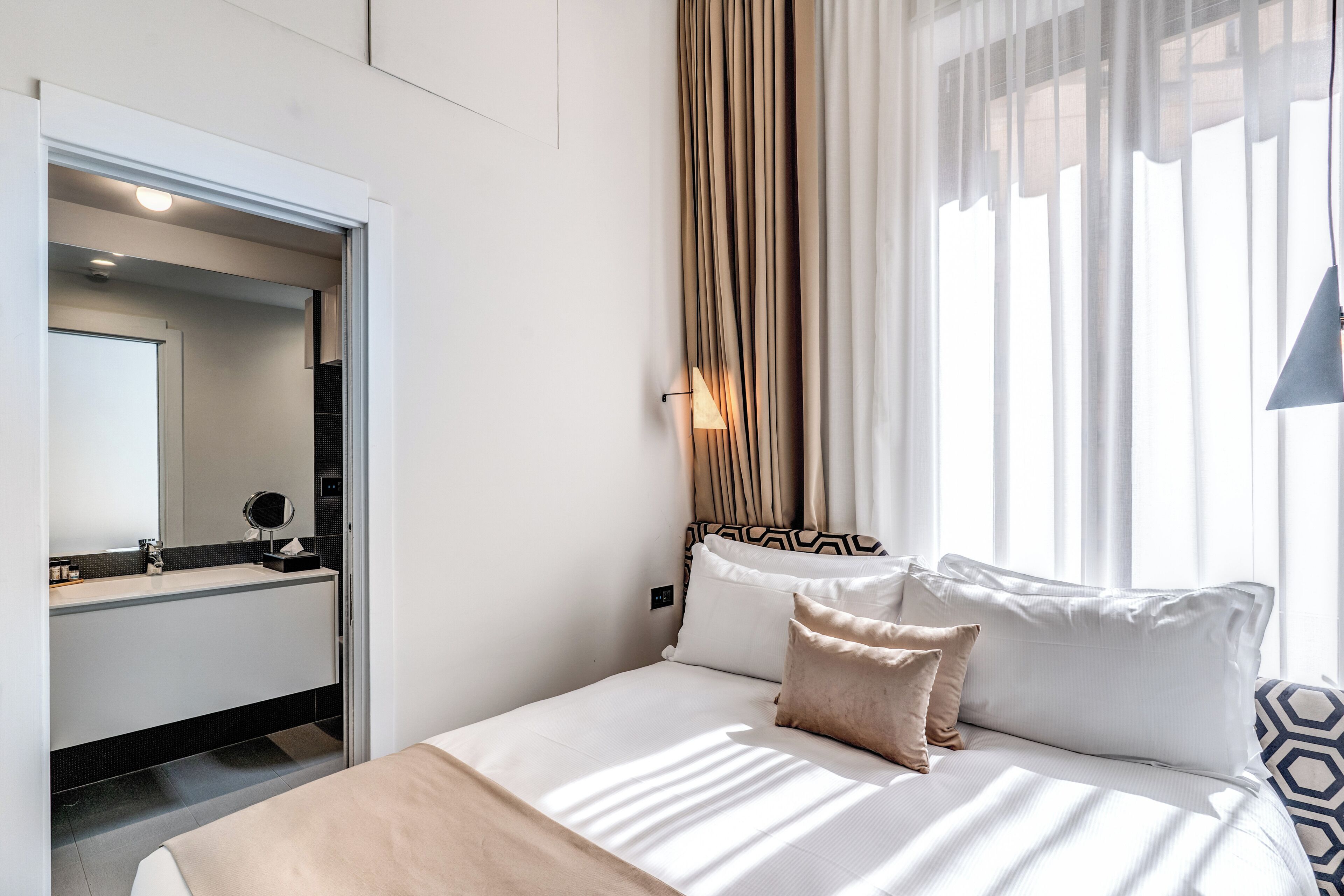 A hotel room with a bed, a bedside table, a mirror, and a window with curtains.