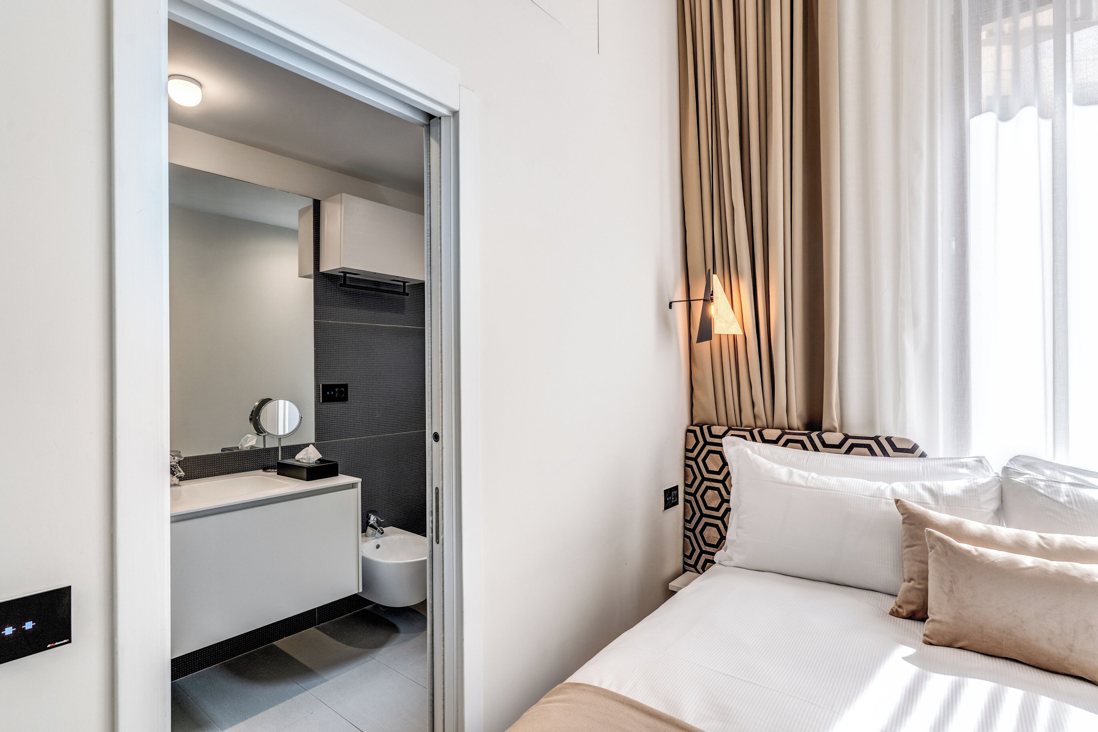 A modern hotel room with a bed, a bedside table, a window with curtains, a bathroom with a sink and mirror, and a door leading to another room.