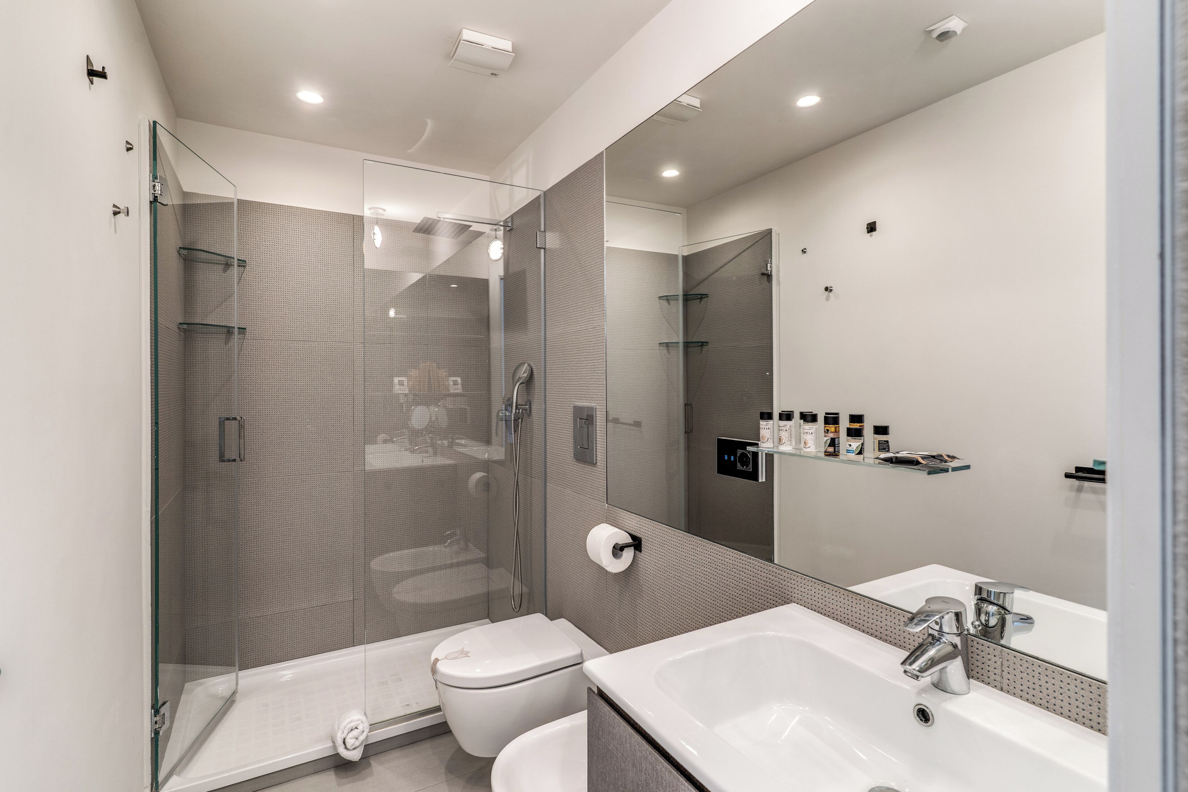 Een moderne badkamer met een douchecabine van glas, een toilet, een wastafel met spiegel en een plank met toiletartikelen.