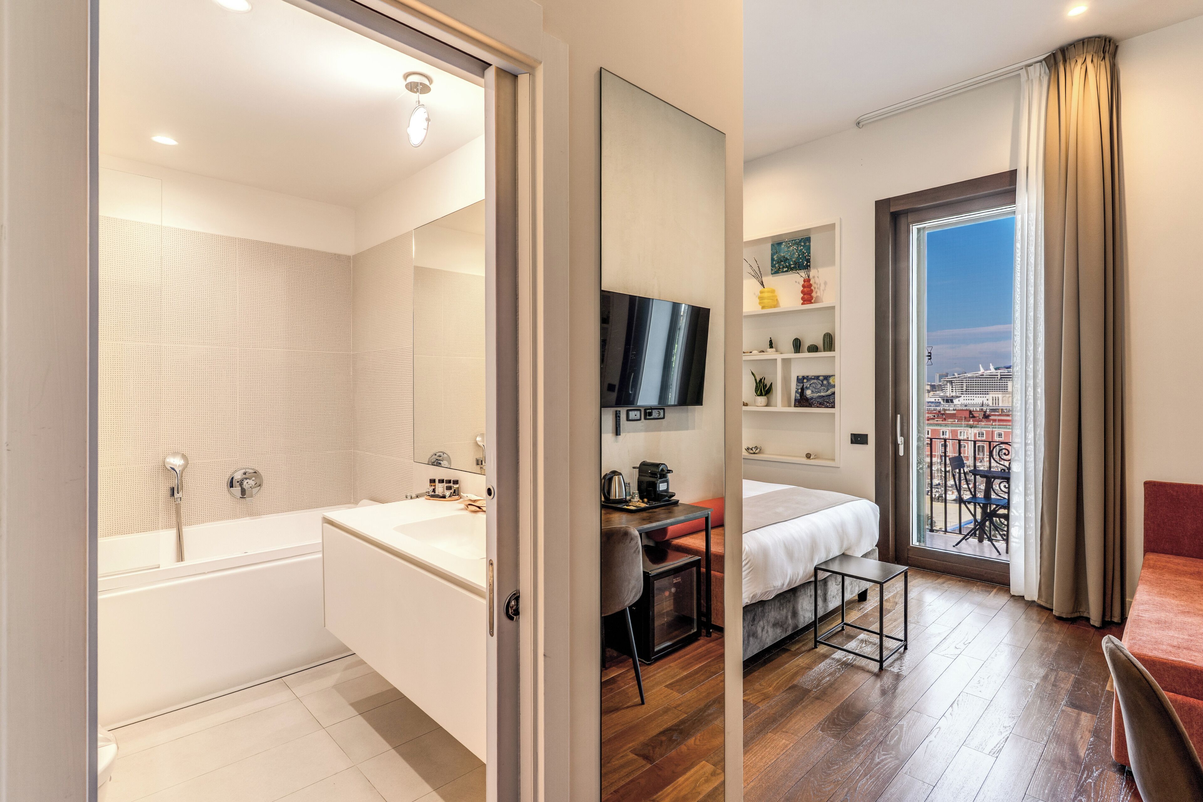 Et moderne hotelværelse med badekar, et skrivebord med stol, seng, fjernsyn og en balkon med byudsigt.