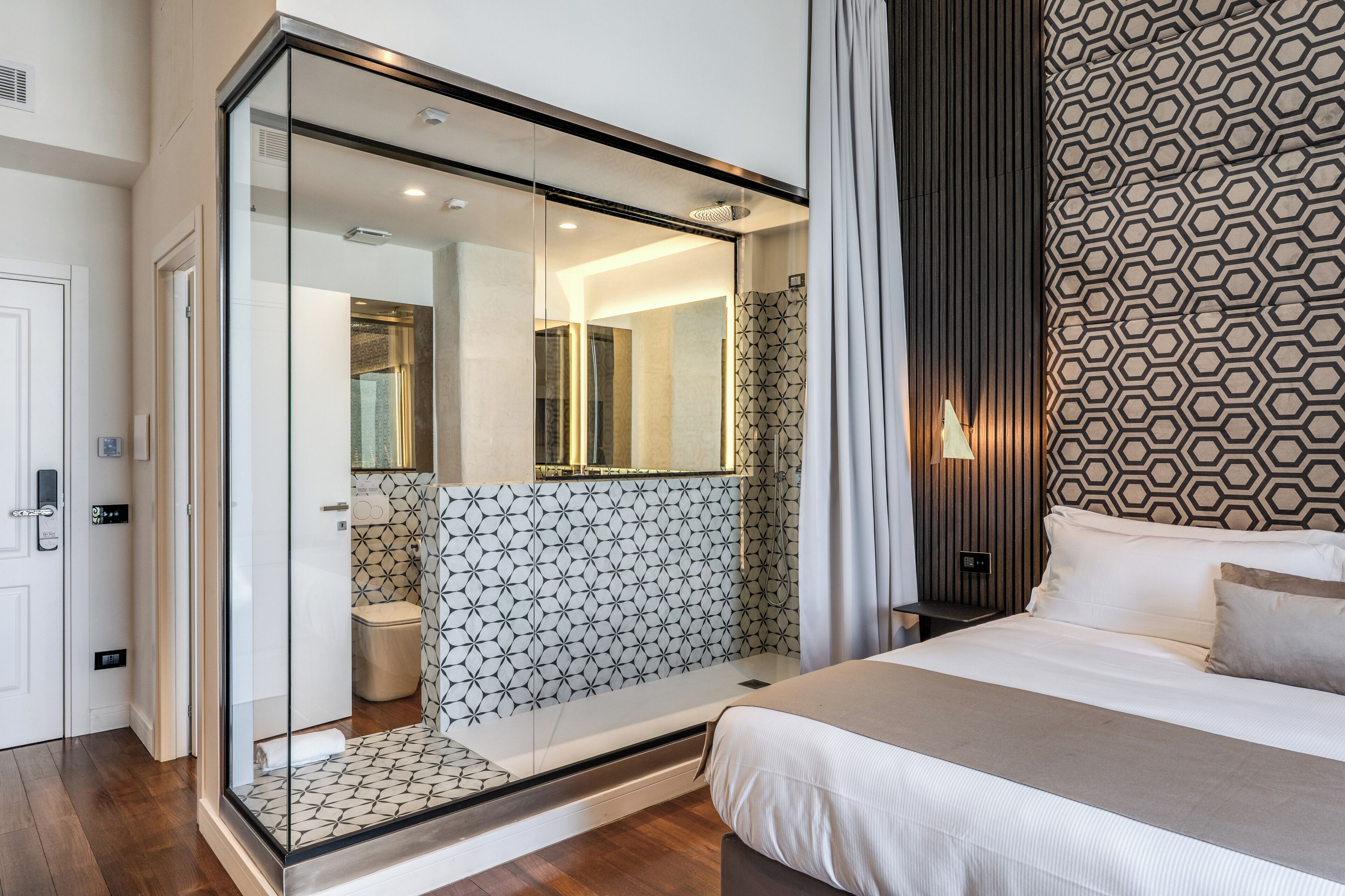 A modern hotel room with a glass-enclosed shower, a bed with a beige bedspread, and a patterned wall.