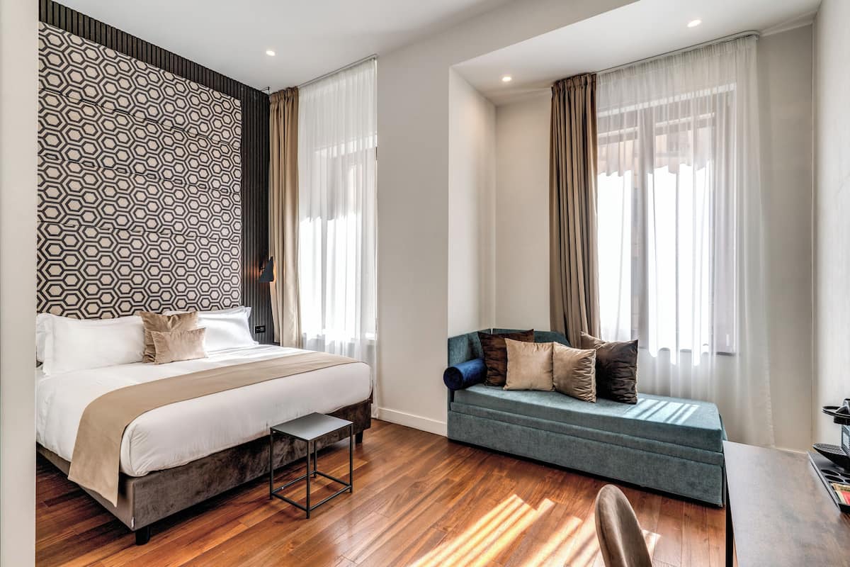 Moderni hotellihuone, jossa on suuri sänky, sohva ja kaksi ikkunaa, joissa on verhot.