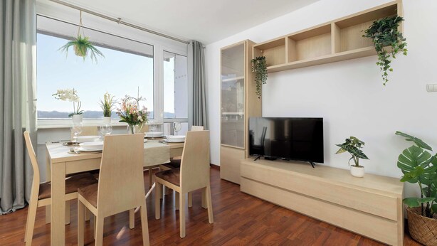 Uma sala de jantar moderna com uma mesa de madeira posta para dois, uma televisão em uma estante de madeira baixa e plantas em vasos.