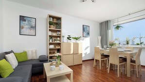 Uma sala de estar moderna com sofá, mesa de jantar e cadeiras. Há uma estante com plantas e objetos decorativos. Uma janela oferece luz natural e vista para o exterior.