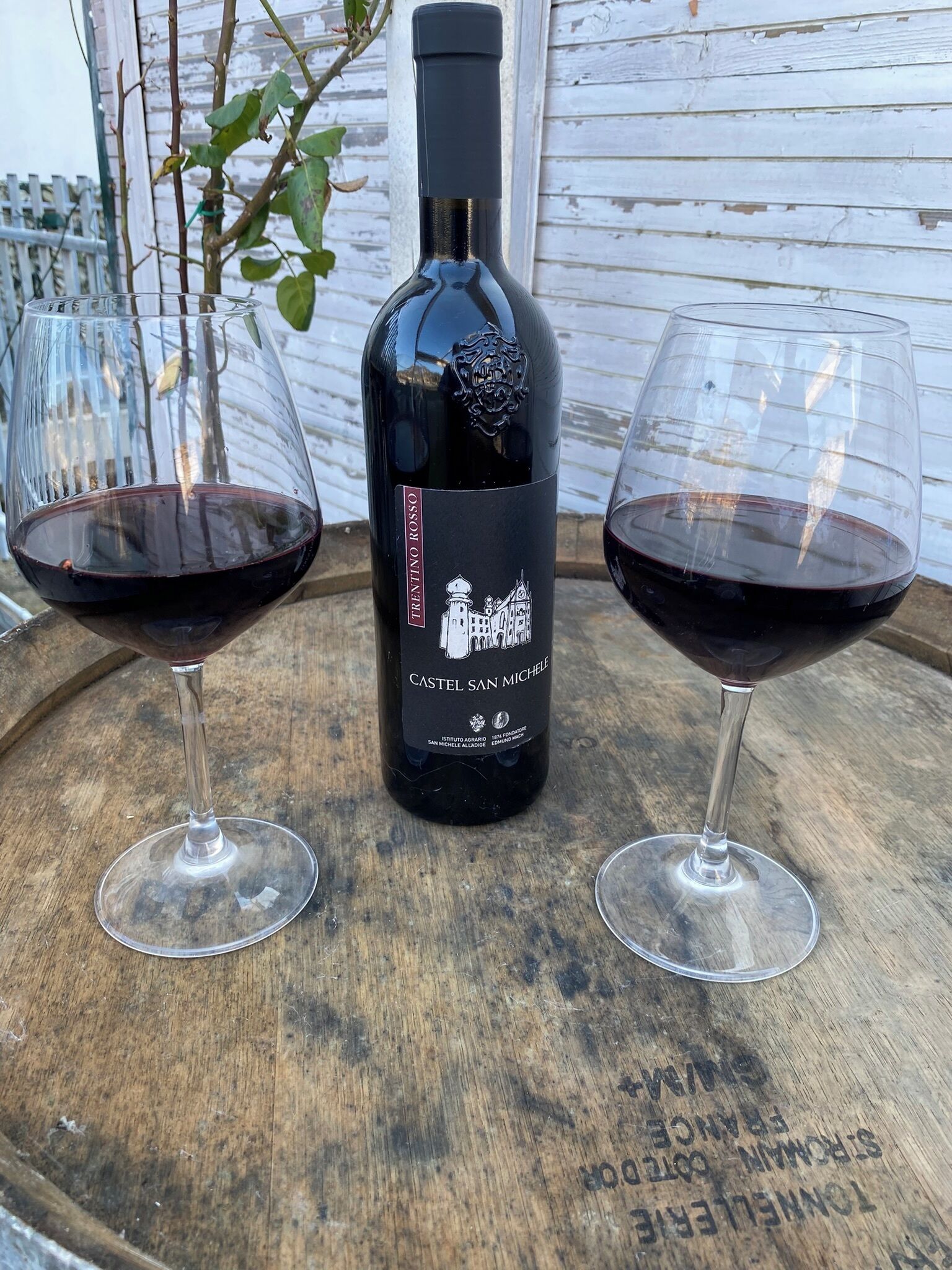 Una botella de vino Trentino Rosso Castel San Michele con dos copas de vino tinto sobre un barril de madera.