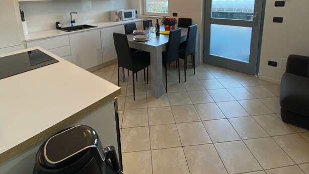 Una cucina moderna con mobili bianchi, un'area pranzo con tavolo e sedie, e un'ampia finestra che offre vista su un edificio.